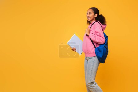 Téléchargez les photos : Adolescente noire joyeuse portant un sac à dos bleu et des classeurs universitaires regardant de côté l'espace de copie, sur fond de studio jaune. Éducation académique offre concept - en image libre de droit