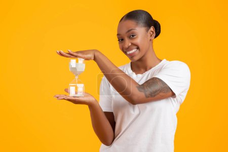 Photo for Time Management. Cheerful African American Woman Holding Sand Clock Smiling To Camera, Posing In White T-Shirt Over Yellow Studio Background. Dont Waste Your Time Concept - Royalty Free Image