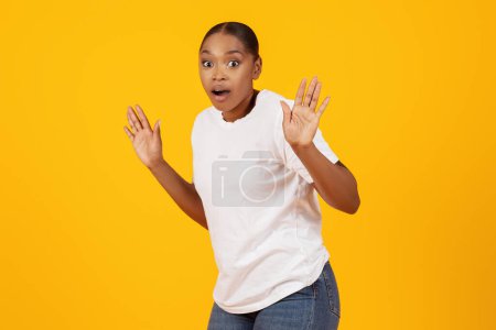 Téléchargez les photos : Confus effrayé dame afro-américaine a été pris posant avec les mains levées en regardant la caméra avec expression effrayée, debout sur fond de studio jaune. Ce n'est pas moi concept - en image libre de droit