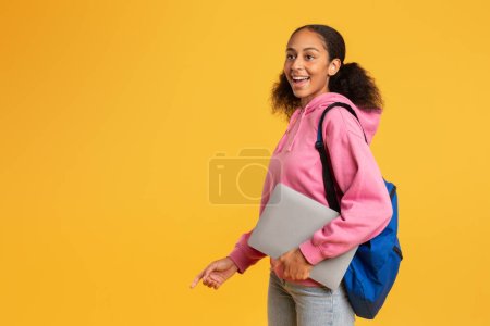 Téléchargez les photos : Étudiante afro-américaine excitée portant son ordinateur portable et son sac à dos sur fond de studio jaune, exprimant son bonheur au sujet du début du semestre d'études. Grande offre éducative. Espace de copie - en image libre de droit