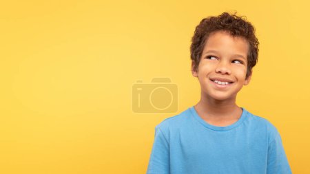 Photo for Contemplative young boy with curly hair looking upward at free space and smiling, dressed in blue shirt against yellow background, suggesting curiosity and imagination - Royalty Free Image