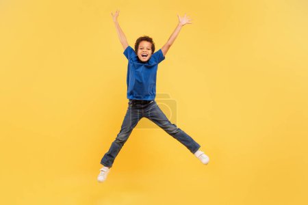 Foto de Niño extático con el pelo rizado saltando alegremente con los brazos extendidos, luciendo camiseta azul y jeans de mezclilla, en un contexto amarillo vivo, encarnando la libertad y la alegría - Imagen libre de derechos