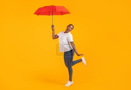 Photo for African american lady in casual posing with red umbrella, enjoying rainy weather in fall or spring, advertising seasonal offer, standing over yellow studio backdrop. Full length, free space - Royalty Free Image
