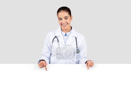 Photo for Radiant european doctor with a stethoscope, smiling and pointing downwards towards an empty banner, ideal for informational text or health advertisement, isolated on gray studio background - Royalty Free Image