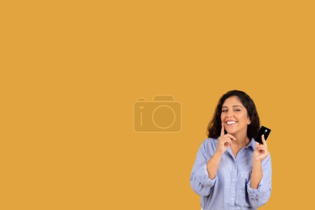 Foto de Mujer joven pensativa con el pelo rizado sosteniendo una tarjeta de crédito, mirando contemplativo, posiblemente considerando una compra o decisión financiera, en una camisa azul sobre un fondo amarillo - Imagen libre de derechos