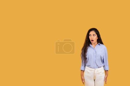Téléchargez les photos : Surprise jeune femme du Moyen-Orient aux longs cheveux ondulés et aux yeux larges, portant une chemise bleue et un pantalon crème, debout avec une bouche ouverte sur un fond jaune uni - en image libre de droit