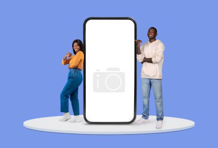 Photo for Smiling young black woman and man pointing and gesturing thumbs up at large blank smartphone screen, suitable for digital adverts, on violet backdrop - Royalty Free Image