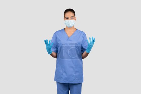 Foto de Una doctora europea compuesta en uniformes azules y guantes protectores levanta las manos, mostrando disposición para un procedimiento estéril o un examen de paciente, aislada sobre fondo gris del estudio - Imagen libre de derechos