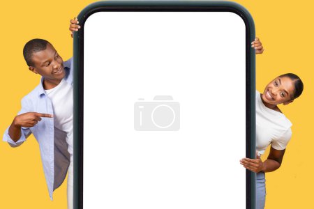 Téléchargez les photos : Homme et femme noirs excités tenant cadre gigantesque smartphone, pointant vers l'écran blanc avec enthousiasme, idéal pour l'application ou l'affichage de la publicité sur fond jaune - en image libre de droit