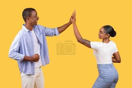 Téléchargez les photos : Black man and woman share playful high-five, leurs visages exprimant joie et camaraderie, sur fond jaune chaud, symbolisant amitié et succès - en image libre de droit