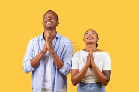Photo for Black man and woman stand with hands clasped in prayer, heads tilted upwards, embodying hope and gratitude on sunny yellow background - Royalty Free Image