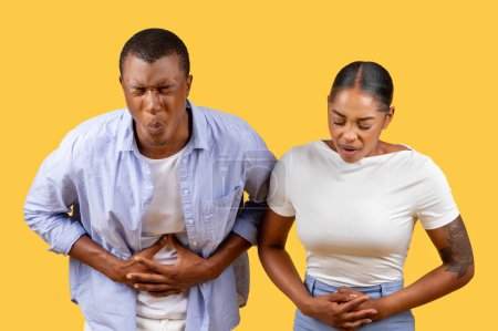 Photo for African american couple clutching their stomachs, faces contorted in discomfort, suggesting digestive upset or pain against plain yellow background - Royalty Free Image