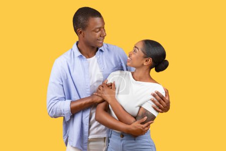 Téléchargez les photos : Joyeux homme et femme noirs dans une étreinte aimante, échangeant des regards tendres et des sourires, rayonnant d'affection sur fond jaune uni - en image libre de droit