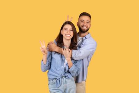 Photo for Laughing woman flashing peace sign with playful man making bunny ears behind her head, both enjoying light-hearted moment on yellow background - Royalty Free Image