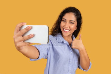 Foto de Jovencita alegre tomando una selfie, dando un gesto de pulgar hacia arriba con su mano libre, sosteniendo el teléfono con una sonrisa brillante, vestida con una camisa azul sobre un fondo amarillo - Imagen libre de derechos