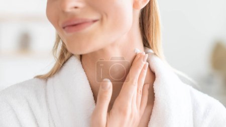Photo for Cropped shot of young blonde lady in bathrobe pampering her neck skin with moisturizer, applying cream and enjoying beauty treatment in modern bathroom setting, closeup portrait - Royalty Free Image