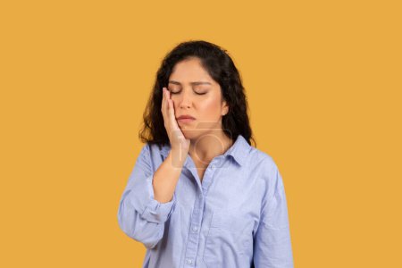 Photo for Woman with a pained expression, hand on her cheek, eyes closed, possibly suffering from a toothache or headache, wearing a light blue shirt against a yellow background, studio - Royalty Free Image