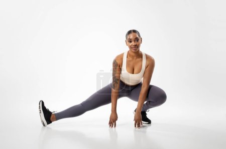 Téléchargez les photos : Femme afro-américaine athlétique exerçant étirement des muscles des jambes dans la posture de squat jambes étendues, ayant entraînement d'entraînement sur fond de studio blanc. Concept de flexibilité. Plan complet - en image libre de droit