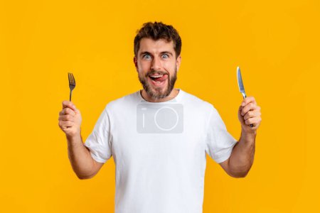 Foto de Joven hambriento sosteniendo cuchillo y tenedor mirando a la cámara, posando sobre fondo amarillo del estudio. Retrato de un joven divertido que tiene antojos de comida y la tentación listo para la cena sabrosa - Imagen libre de derechos