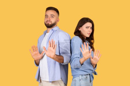 Téléchargez les photos : Homme et femme montrant les paumes en avant en arrêtant le geste, avec des expressions méfiantes suggérant prudence ou refus, sur fond jaune propre - en image libre de droit
