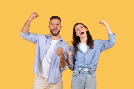 Foto de Exuberante pareja europea joven celebrando la victoria con los puños levantados y sonrisas anchas, exudando felicidad y éxito contra el fondo amarillo brillante - Imagen libre de derechos