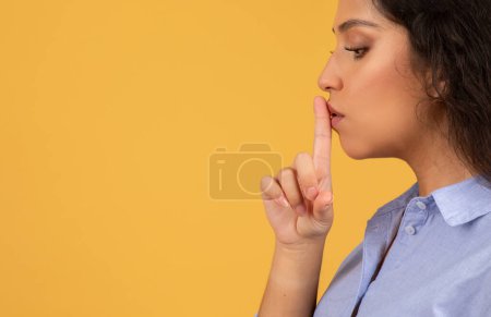 Foto de Perfil lateral de una joven mujer de Oriente Medio con el pelo rizado colocando su dedo índice sobre los labios en un gesto shh, señalando silencio o secreto, sobre un fondo amarillo - Imagen libre de derechos