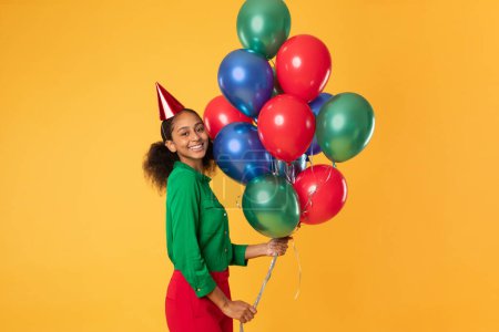 Téléchargez les photos : Joyeux adolescent noir fille en chapeau d'anniversaire tenant un tas de ballons colorés, souriant à la caméra, posant sur fond de studio jaune. Bday célébration et concept de fête - en image libre de droit