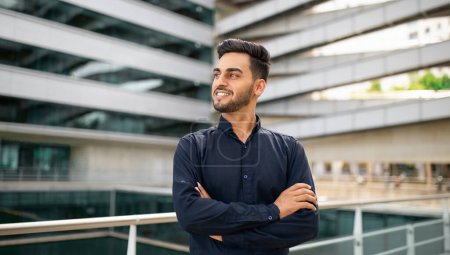 Téléchargez les photos : Un jeune homme d'affaires souriant du Moyen-Orient en chemise noire élégante croise les bras en toute confiance sur fond d'immeuble de bureaux moderne dans la région de la ville, exsudant succès et professionnalisme - en image libre de droit