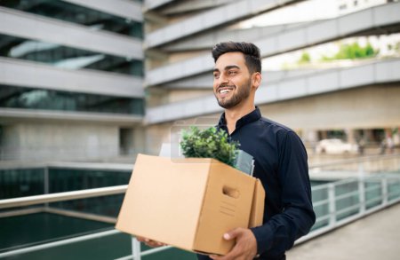 Photo for New career start. Hopeful young professional arabic businessman carries belongings box in hands, begins job transition, reflecting optimism amidst change, leaving office building - Royalty Free Image