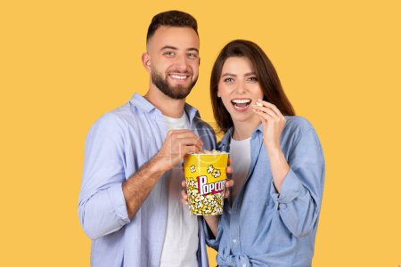Photo for Happy couple shares bucket of popcorn, the man and woman both smiling joyfully, suggesting cozy movie night in, against cheerful yellow background - Royalty Free Image