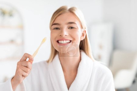Foto de Mujer rubia europea manteniendo la salud bucal cepillándose los dientes, usando pasta de dientes para blanquear, disfrutando de parte de su cuidado diario en el moderno baño doméstico, sonriendo a la cámara - Imagen libre de derechos