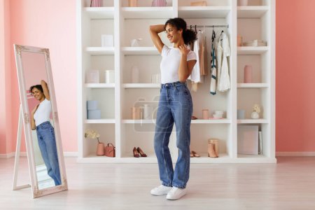 Photo for Smiling latin woman feeling confident in her high-waisted jeans, striking pose while looking at her reflection in full-length mirror in pink room - Royalty Free Image