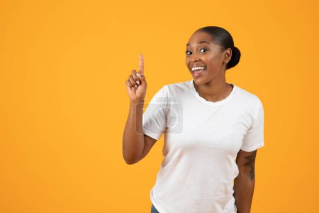Photo for Happy young black lady in t-shirt pointing finger upwards on yellow studio background. Millennial African American woman experiencing AHA eureka moment, having creative idea, empty space - Royalty Free Image