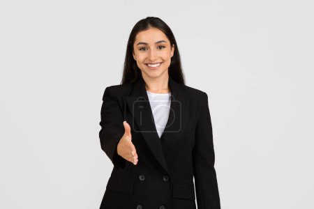Téléchargez les photos : Heureuse jeune femme d'affaires européenne qui tend la poignée de main avec un sourire accueillant, signe de l'affaire, posant dans un élégant costume noir sur fond de studio gris - en image libre de droit