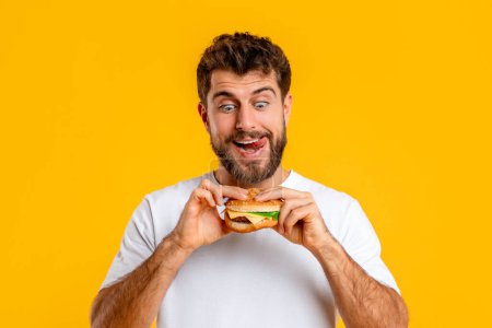 Téléchargez les photos : Joyeux jeune homme affamé mangeant un hamburger posant sur fond jaune, Studio Shot. Mangeur de malbouffe Profiter d'une nutrition déséquilibrée Savourer un cheeseburger malsain. Concept de repas Cheat - en image libre de droit