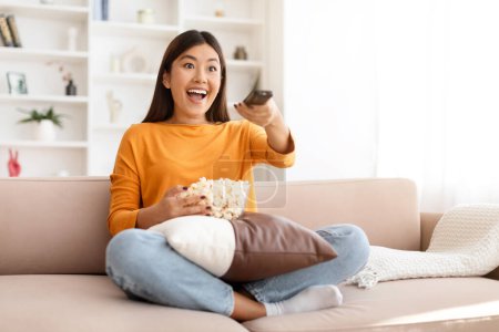 Téléchargez les photos : Jeune femme asiatique excitée regardant son émission de télévision préférée, assise sur le canapé dans le salon, tenant bol avec pop-corn et télécommande, profitant week-end devant la télévision, espace de copie - en image libre de droit