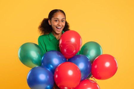 Foto de Feliz chica adolescente afroamericana sostiene racimo de globos festivos de colores sobre fondo de estudio amarillo, expresando alegría de fiesta de cumpleaños y celebración de vacaciones, sonriendo a la cámara - Imagen libre de derechos