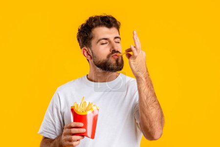 Photo for Junk Food. Portrait Of Hungry Man Eating Tasty French Fries Standing Over Yellow Studio Background, Enjoying Taste Of Unhealthy Snack. Concept Of Cheat Meal, Unbalanced Nutrition - Royalty Free Image