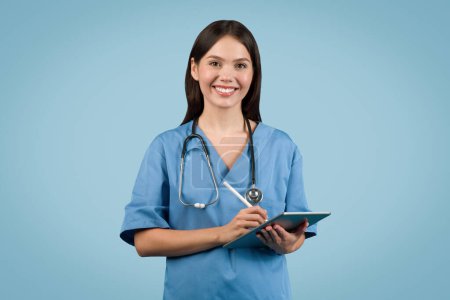 Téléchargez les photos : Joyeux jeune infirmière en manteau bleu tenant tablette numérique et stylet, offrant un sourire amical à l'appareil photo, sur fond bleu - en image libre de droit