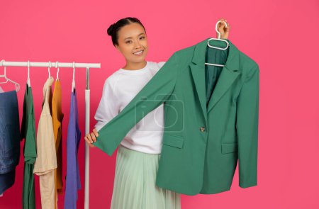 Foto de Mujer asiática alegre demostrando ropa nueva mientras está de pie junto a estante de moda, mujer coreana sonriente sosteniendo chaqueta chaqueta de moda en la mano, posando aislado sobre fondo de estudio rosa - Imagen libre de derechos