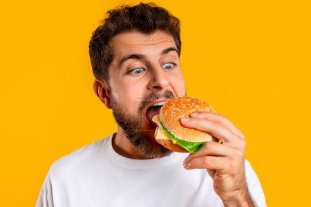 Photo for Closeup Portrait Of Hungry Young Guy Eating Burger In Studio On Yellow Background, Biting Tasty Cheeseburger Snacking With Junk Food. Unbalanced Nutrition, Cheat Meal Concept - Royalty Free Image