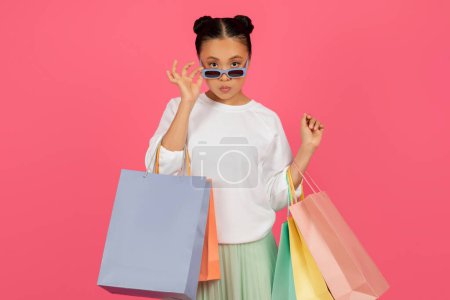 Foto de Mujer asiática con estilo con bolsas de compras ajusta sus gafas de sol y mirando a la cámara, joven coreana shopaholic dama llevando compras, disfrutando de la moda y las ventas, de pie aislado sobre fondo rosa - Imagen libre de derechos