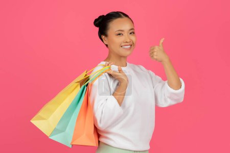 Foto de Gran concepto de compras. Retrato de la señora asiática feliz sosteniendo coloridas bolsas de comprador y mostrando el gesto de pulgar hacia arriba, mujer alegre mirando a la cámara, de pie aislado en el fondo rosa - Imagen libre de derechos
