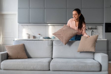 Photo for Engaged woman carefully arranging and fluffing throw pillows on sofa, taking pride in her neat and stylishly decorated living space with modern kitchen backdrop - Royalty Free Image