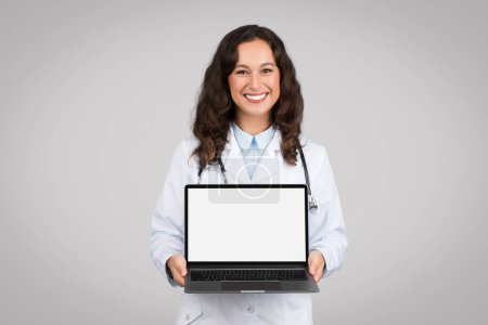 Téléchargez les photos : Heureux médecin femme montrant ordinateur portable avec écran vide publicité offre médicale en ligne ou un service debout sur fond de studio gris, souriant à la caméra. Maquette - en image libre de droit