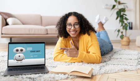 Téléchargez les photos : Joyeux afro-américain jeune femme portant des lunettes étudiant couché sur le tapis à la maison, en utilisant chat bot sur ordinateur portable, pointant vers l'écran, faire des devoirs, livre de lecture, collage - en image libre de droit