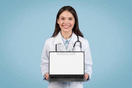 Téléchargez les photos : Joyeux médecin féminin présentant un ordinateur portable avec écran vierge, symbolisant la facilité d'accès aux services de e-santé et à la publicité médicale - en image libre de droit
