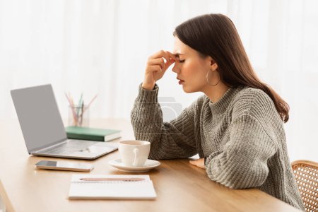 Photo for Exhausted young woman entrepreneur sitting at desk with laptop, cup of coffee and stationery on side, touching her nose bridge, suffer from sore eyes, home office interior. Burnout, overworking - Royalty Free Image