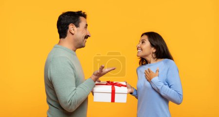 Téléchargez les photos : Un homme excité dans un pull vert présente une boîte cadeau avec un ruban rouge à une femme surprise dans un haut bleu, tous deux engagés avec des expressions joyeuses sur un fond orange vif - en image libre de droit