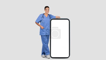 Foto de Sonriente enfermera médica milenaria europea en uniforme azul con gran teléfono inteligente con pantalla en blanco, aislado sobre fondo gris, longitud completa, panorama. Recomendación médica en línea, aplicación de atención médica - Imagen libre de derechos